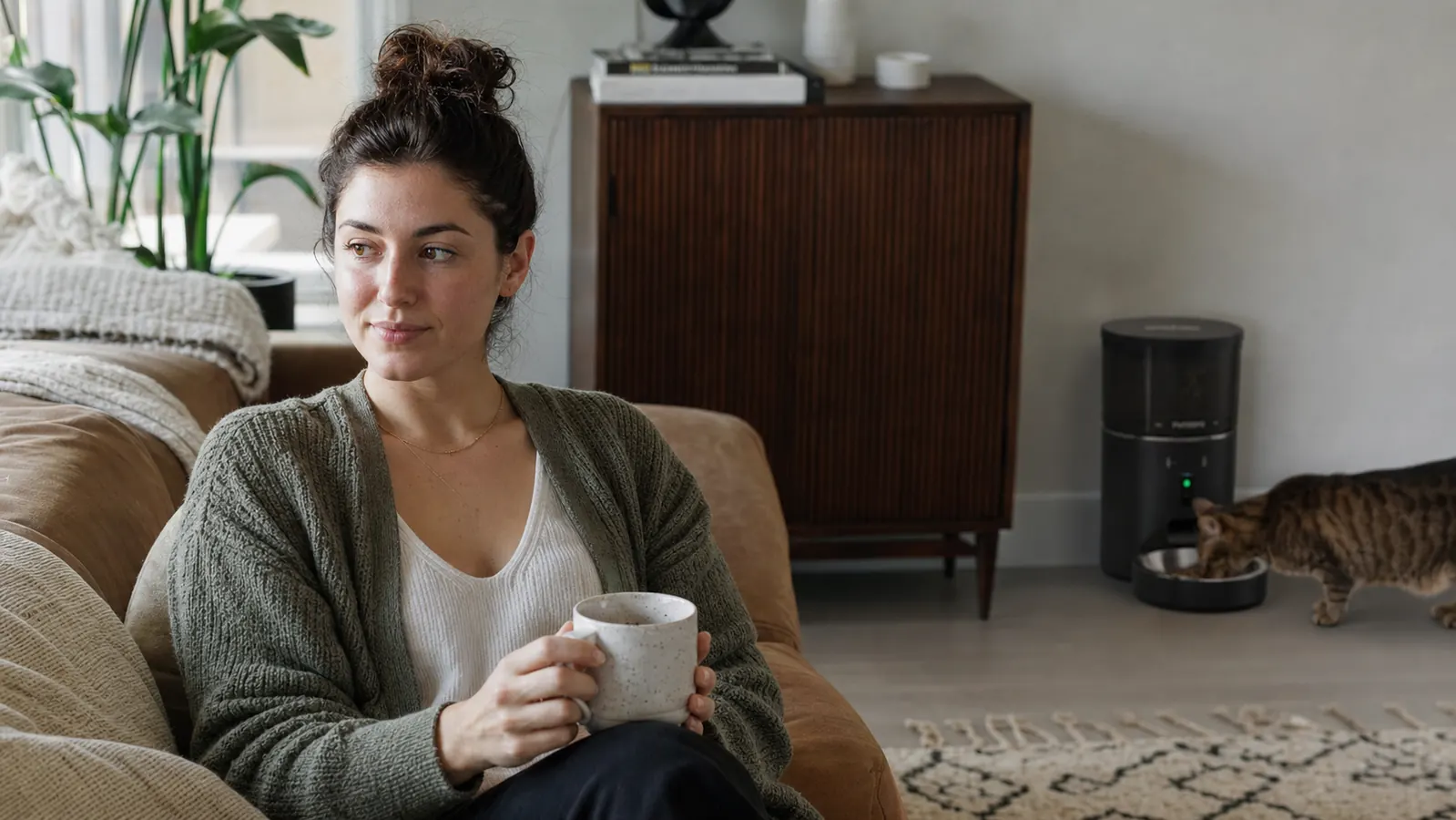 Person relaxing on a couch with a coffee mug while a cat eats from an automatic feeder in the background
